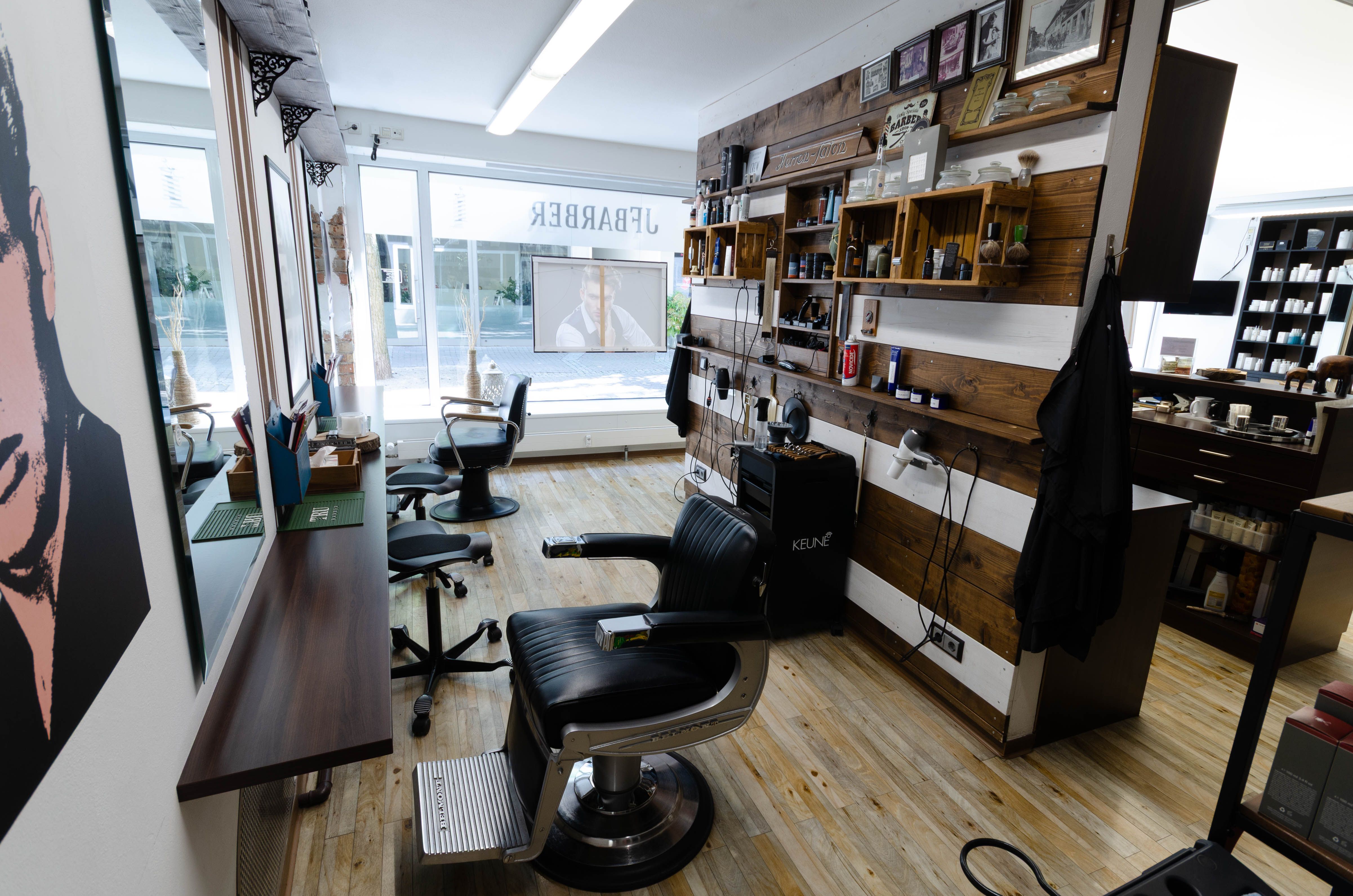 Blick durch den Barber-Bereich mit einem klassischen schwarzen Barber-Stuhl, einem Spiegel mit schmaler Holzkonsole und einem Pop-Art-Porträt an holzverkleideten Wänden mit Pflegeprodukten, dahinter die Glastür zum Eingang