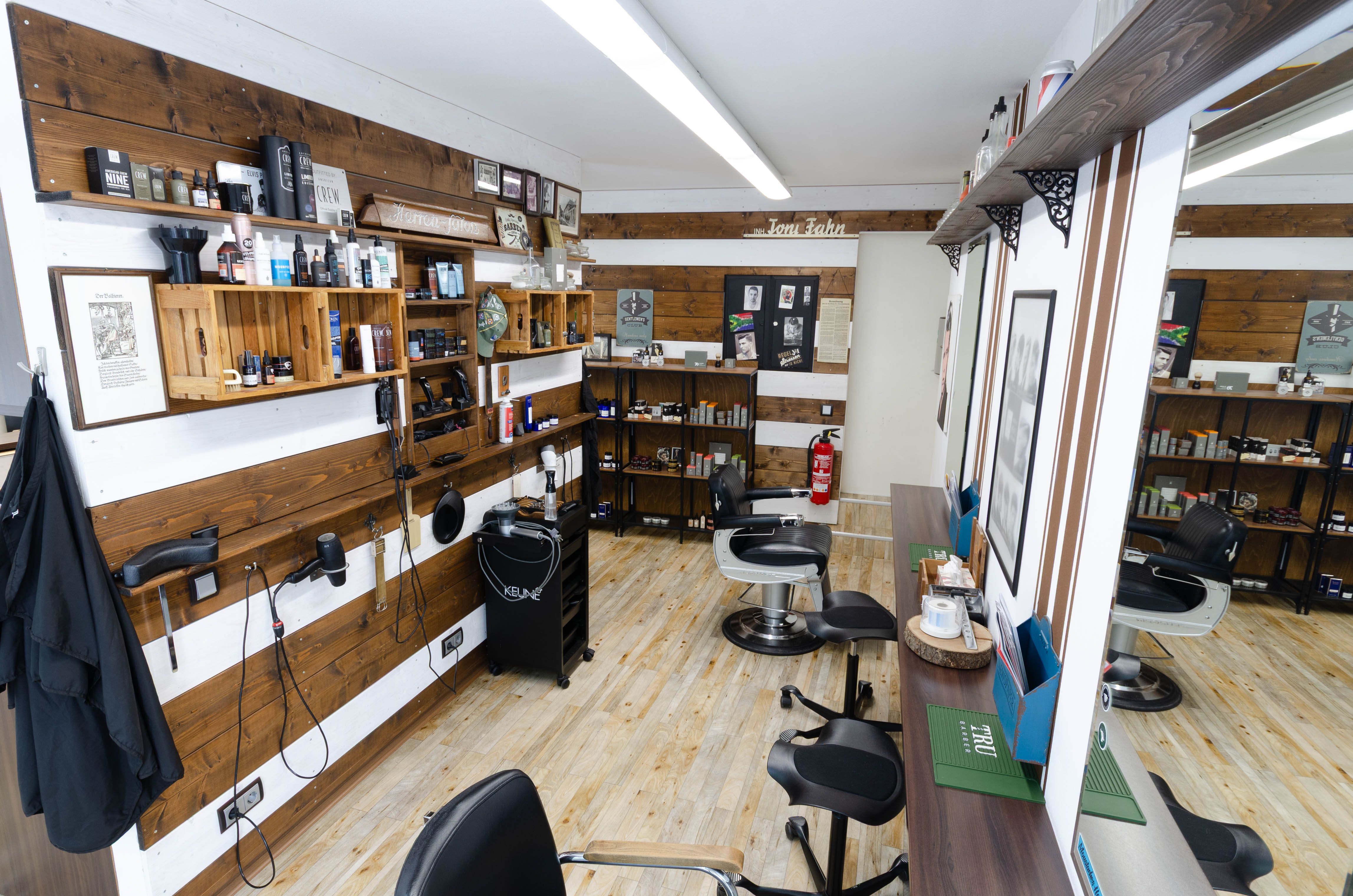 Blick in den Barber-Bereich mit zwei klassischen Barber-Stühlen, holzverkleideten Wänden voller Pflegeprodukte, Föhnen und Spiegeln